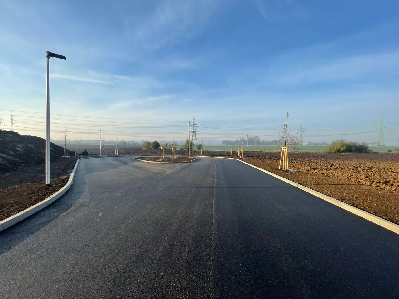 Nově vyasfaltovaná silnice s několika mladými stromy zasazenými podél okrajů a pouličním osvětlením, na pozadí zemědělská pole a modrá obloha.
