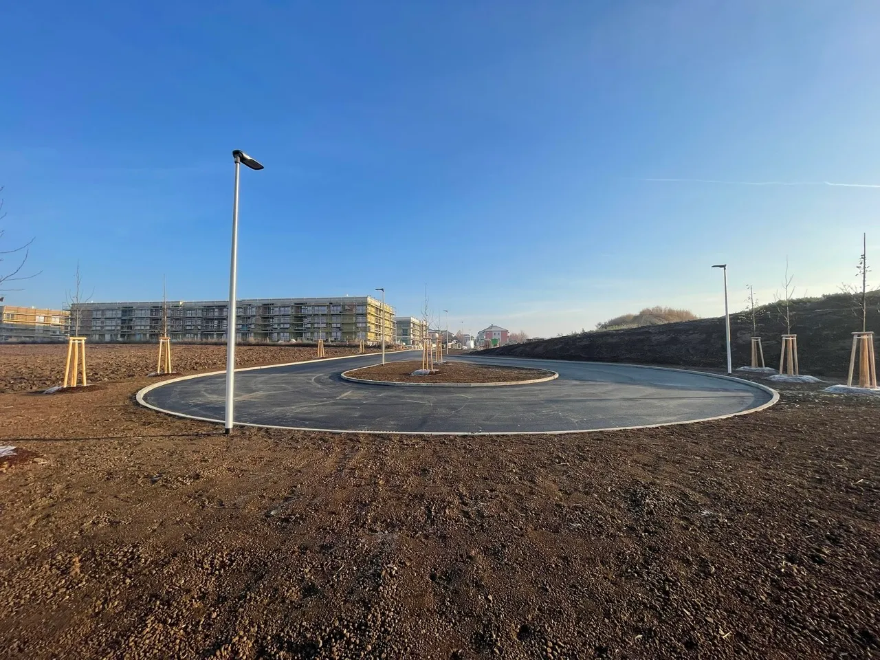 Nově vyasfaltovaný kruhový objezd obklopený holou zemí a mláděcími stromy s budovami v pozadí pod modrou oblohou.