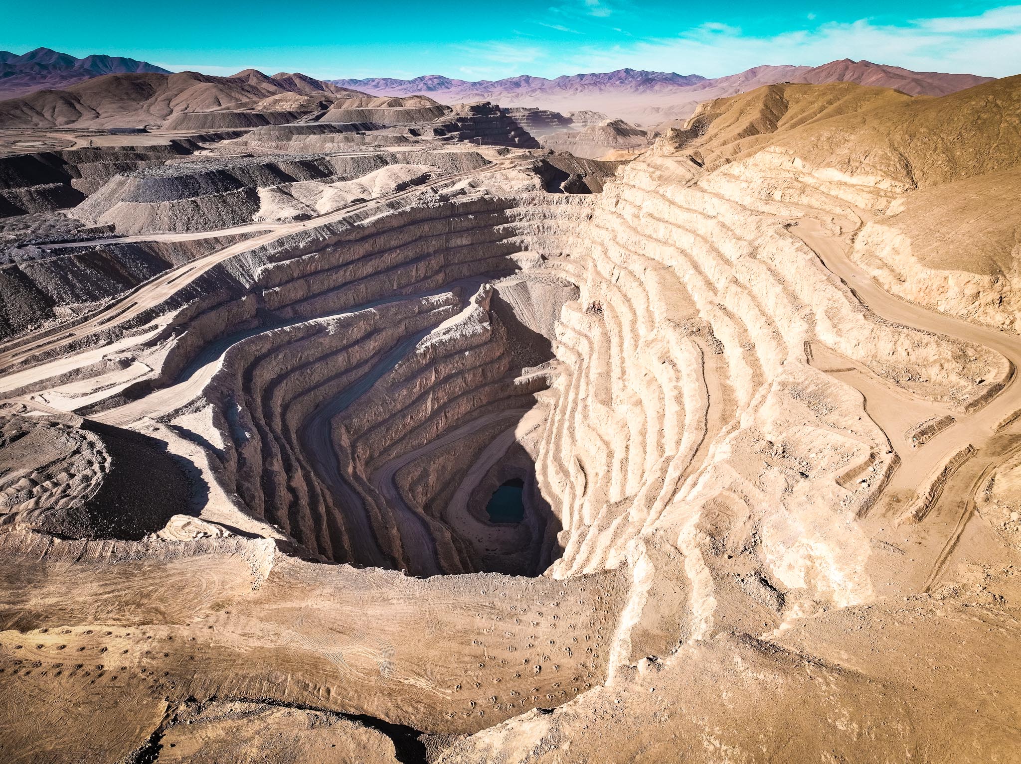 Aerial view of a large open-pit mine with terraced layers carved into the earth and mountainous terrain in the background.