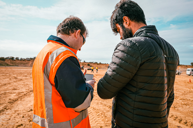 Two men standing on a mine site, one wearing an orange safety vest and the other a black puffer jacket, both looking at a smartphone.