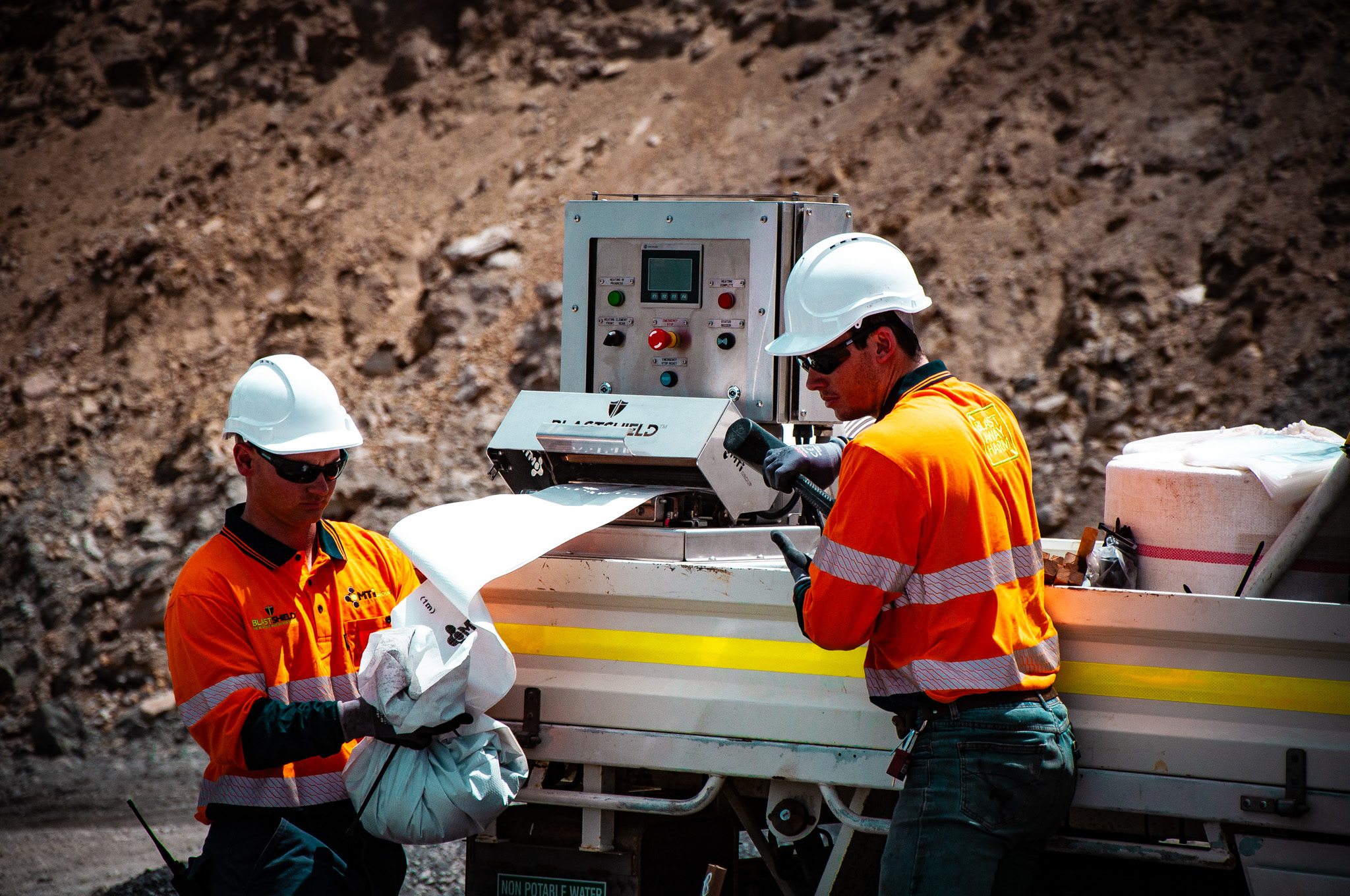 Two workers in orange safety jackets and white helmets operating the Blastshield hole lining machine outdoors near a rocky terrain.