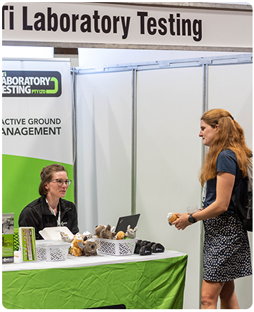 Woman with backpack interacting with a seated staff member at an MTi Laboratory Testing exhibit booth.