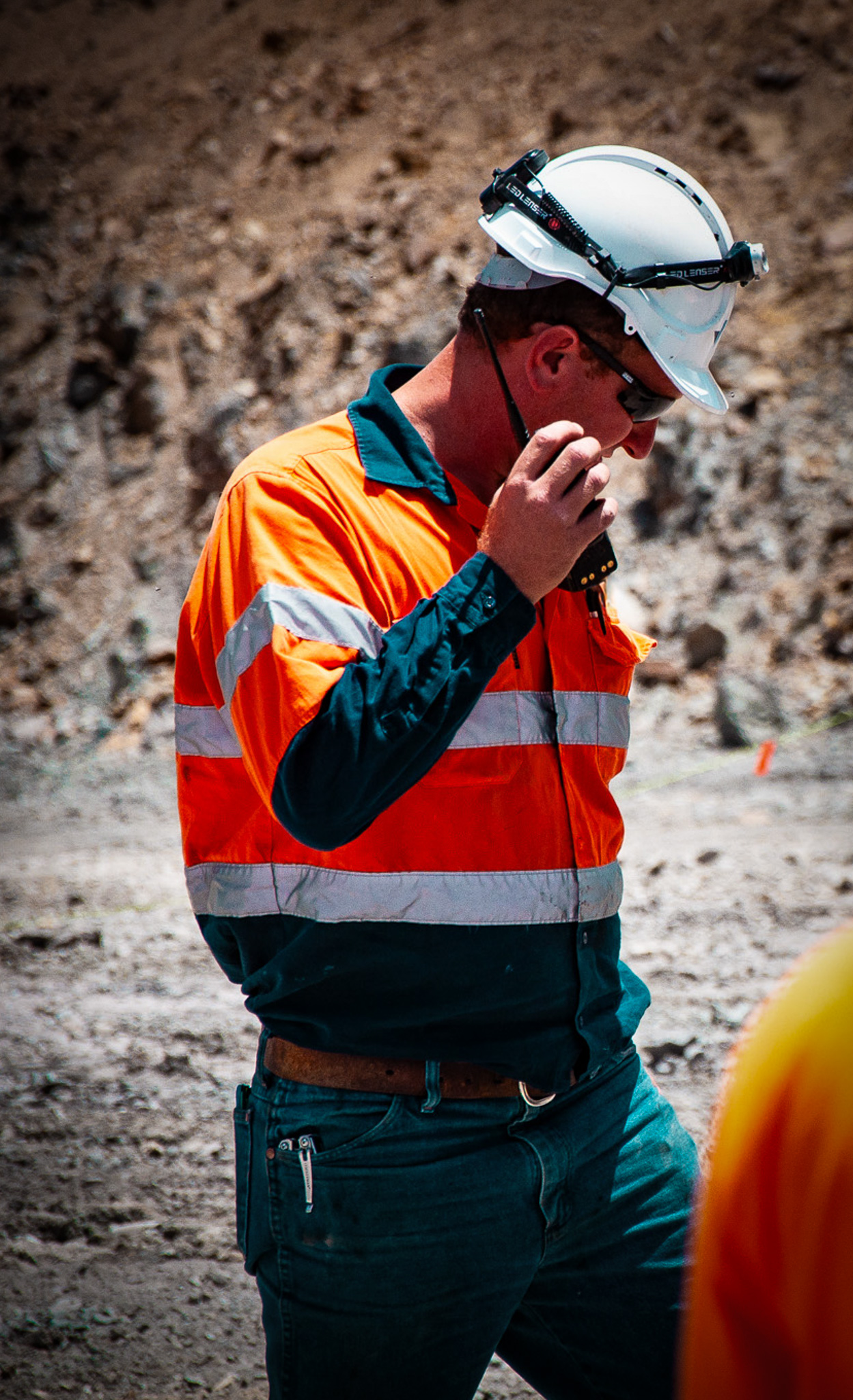 Mine worker in an orange and navy reflective jacket and white hard hat speaking into a walkie-talkie at a rocky outdoor site.