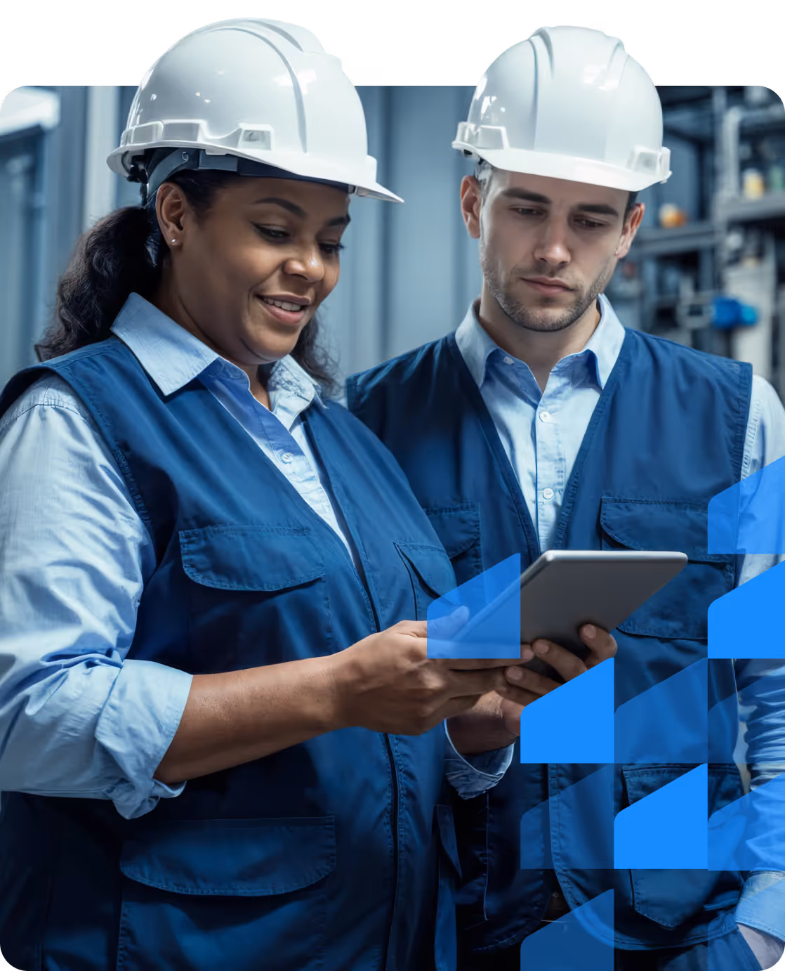 Two engineers wearing white hard hats and blue vests looking at a digital tablet in an industrial setting.