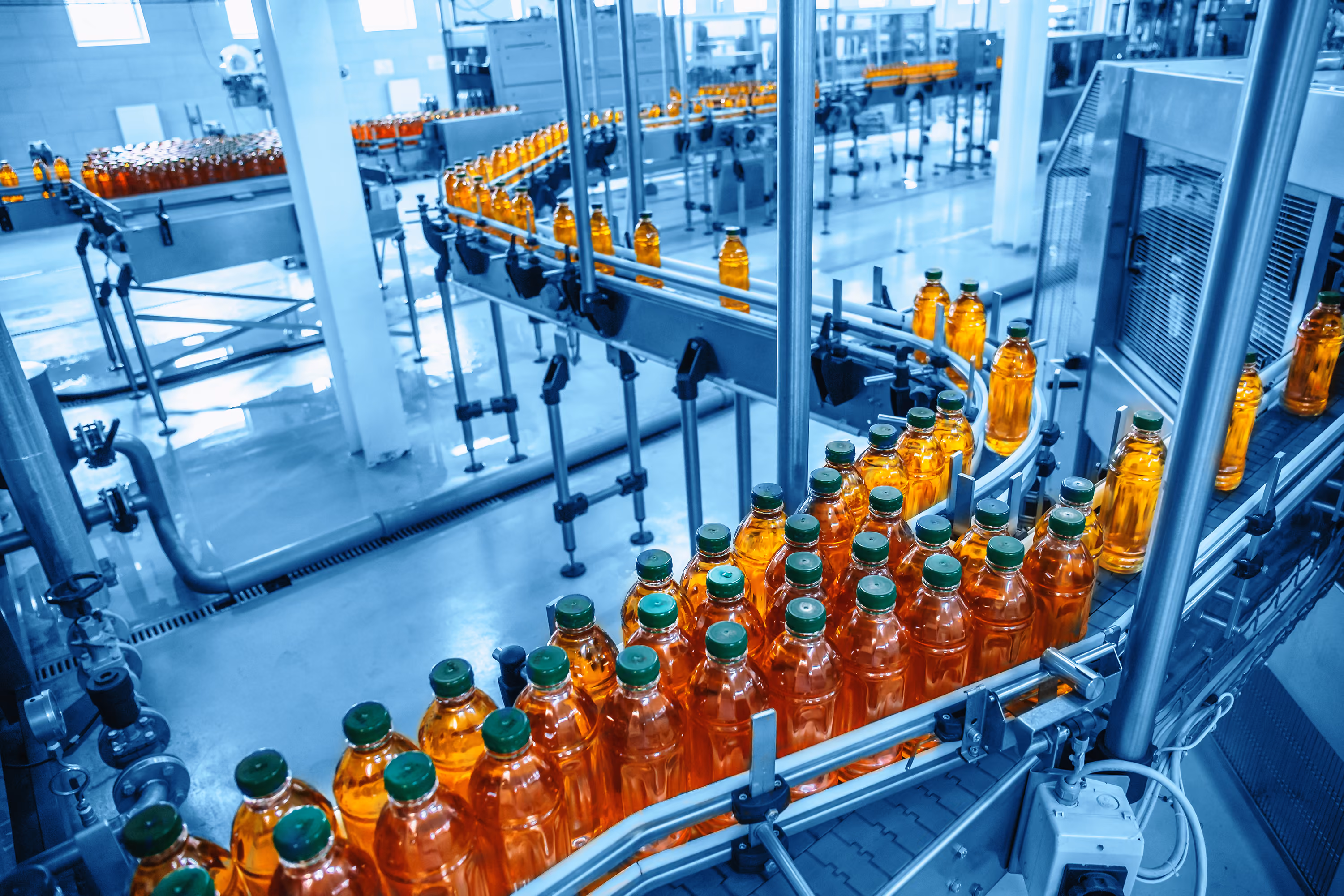Bottling factory conveyor system transporting rows of filled transparent orange bottles with green caps through the production line.