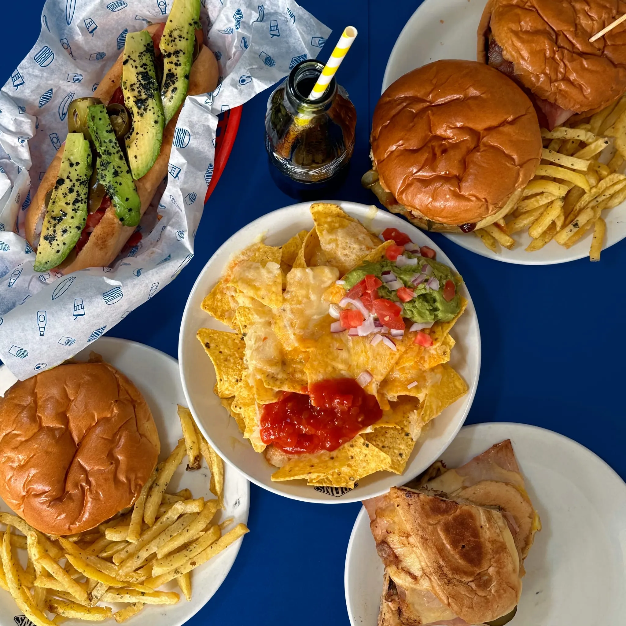 Varias hamburguesas con papas fritas, un sándwich, nachos con queso, guacamole, salsa y un hot dog con aguacate en una mesa azul.