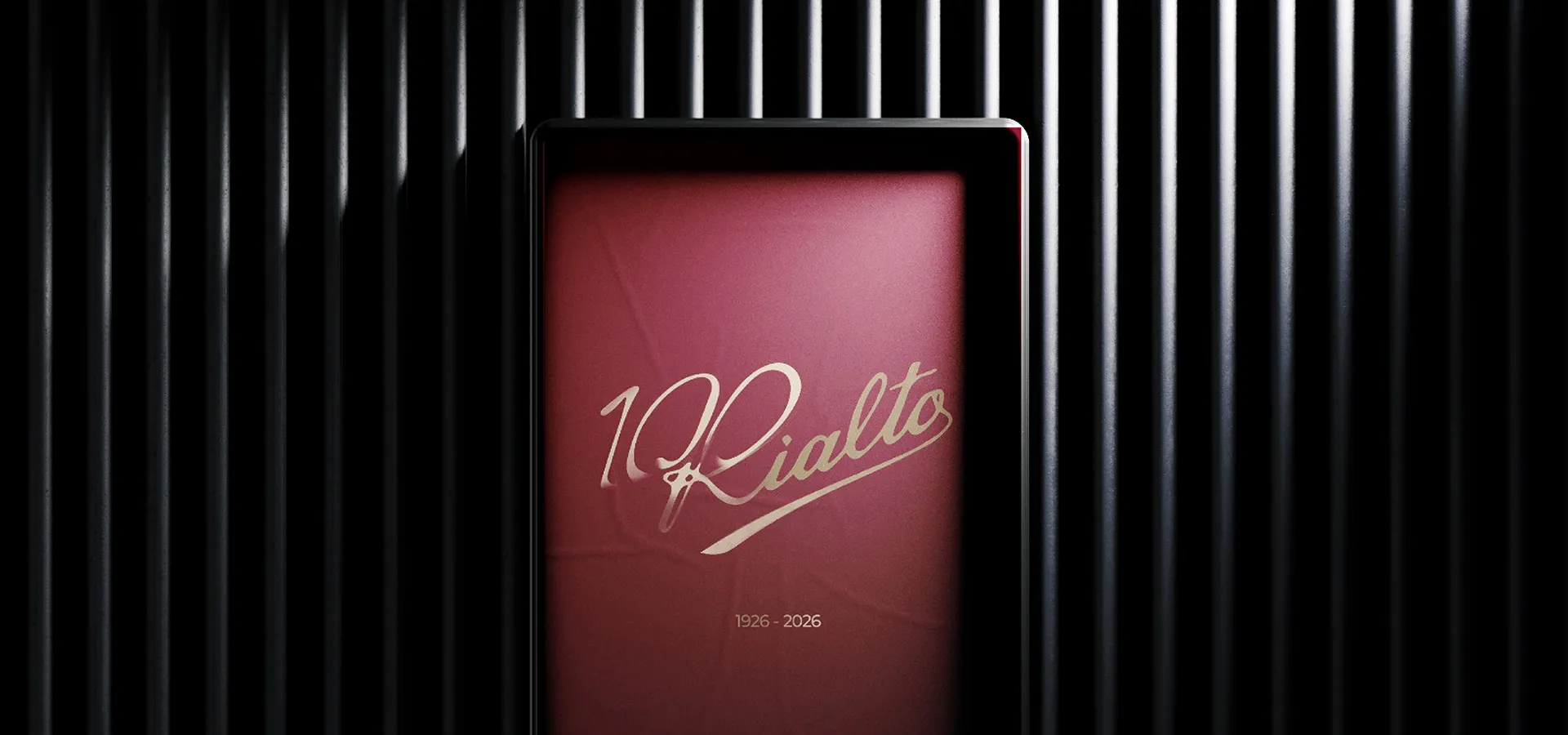 Cartel rojo con texto dorado que dice '100 Rialto' y las fechas 1926 - 2026 sobre fondo de listones verticales negros.