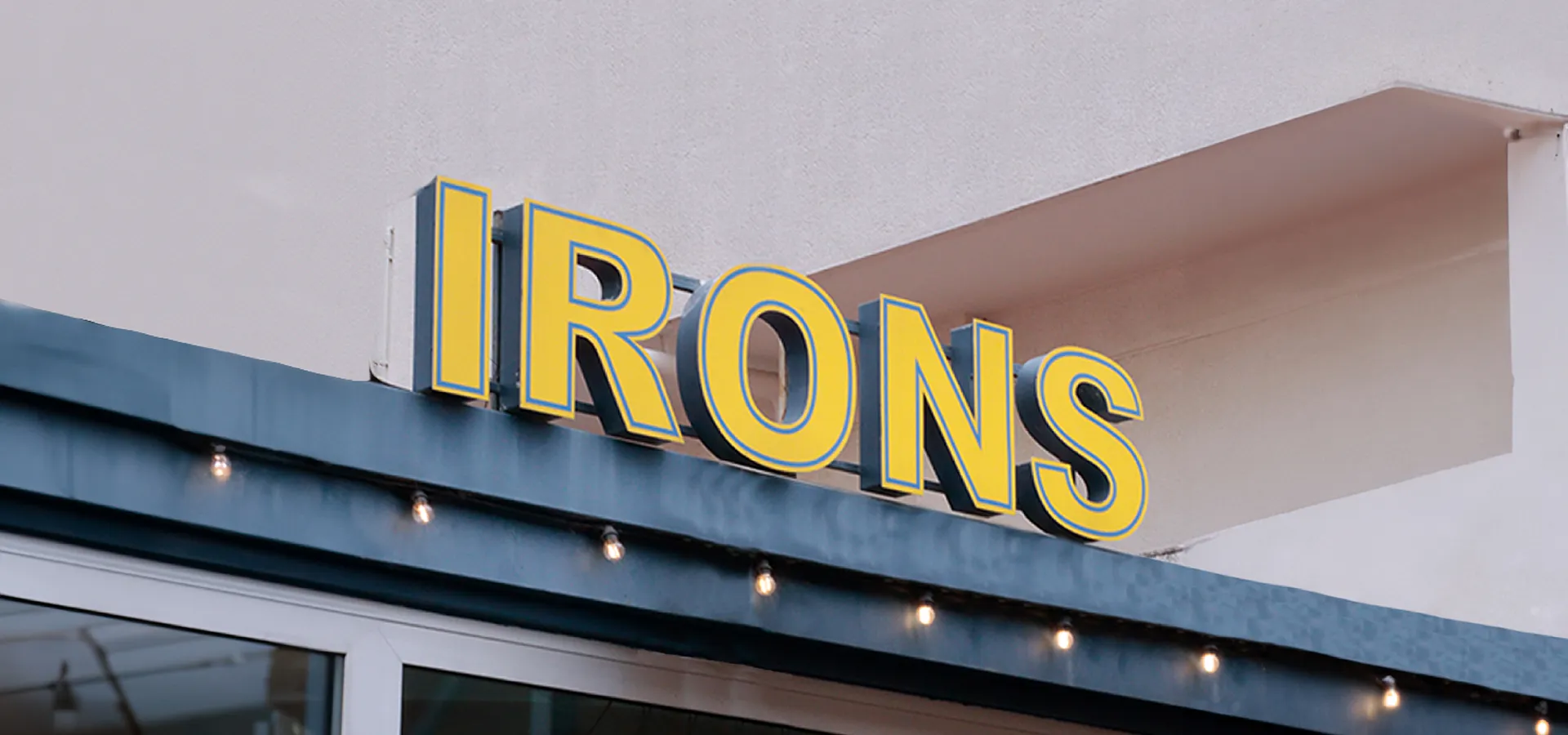 Letrero amarillo con borde azul que dice IRONS sobre una estructura con luces colgantes.