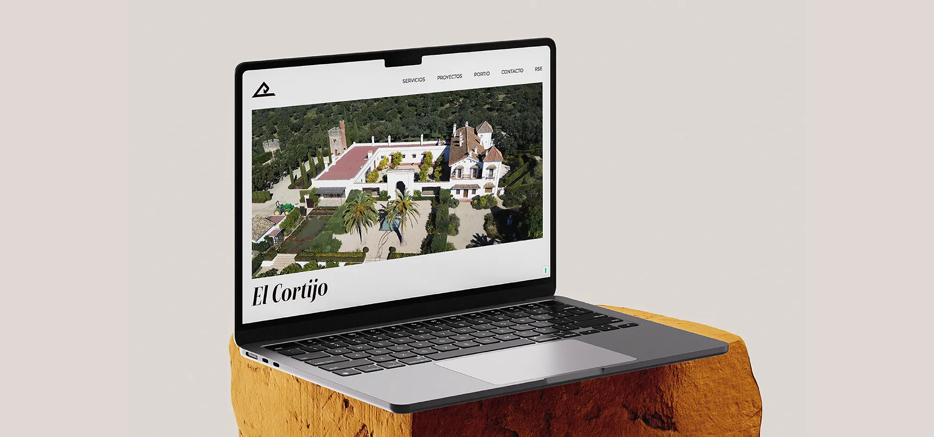 Portátil abierto sobre una roca amarilla que muestra una página web con una imagen aérea de una casa grande y jardines titulada El Cortijo.