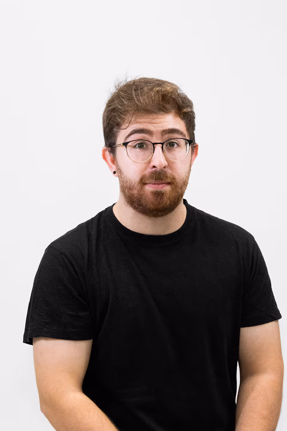 Joven con gafas, barba y camiseta negra mirando hacia adelante sobre fondo blanco.