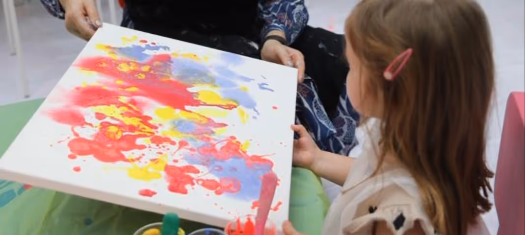A girl watching an adult tilt a canvas to spread vibrant red, yellow, and blue paint.