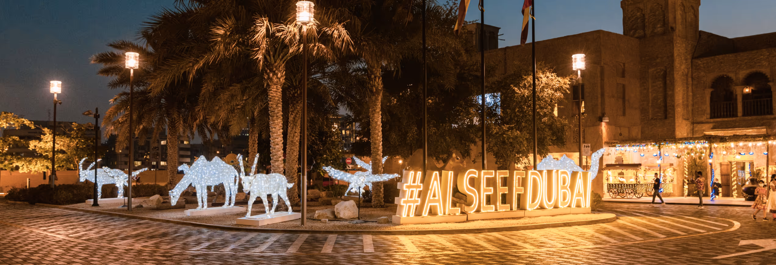 Glowing #ALSEEFDUBAI sign and animal light sculptures in a palm-lined plaza at night