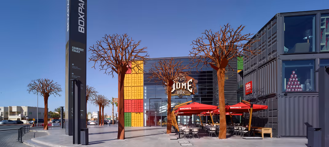 Modern urban space featuring "BOXPARK" signage, container-style buildings, and metal tree art.