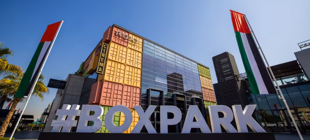 Modern urban space featuring "BOXPARK" signage, container-style buildings, and metal tree art.