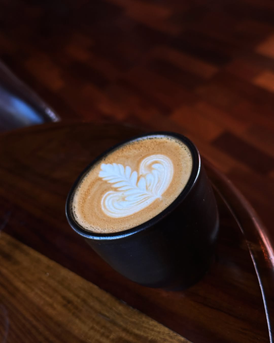 A small black cup of latte featuring a crisp white leaf and heart latte art on a wooden table.