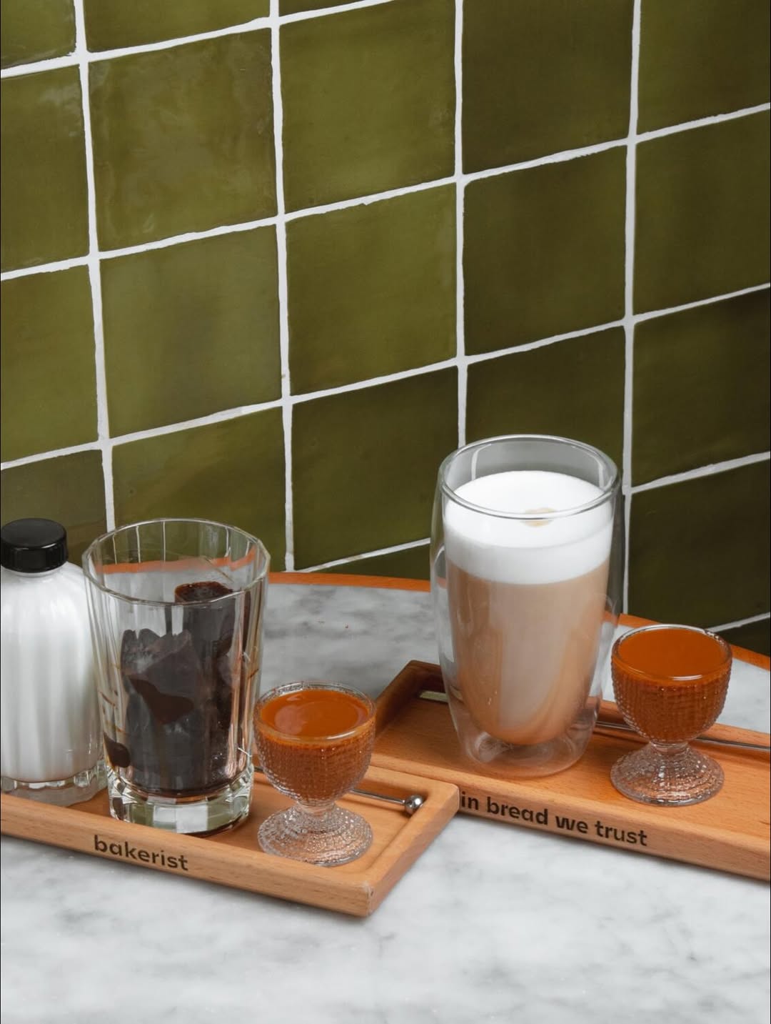 Various coffee drinks and ingredients served on "bakerist" wooden trays against green tiles.