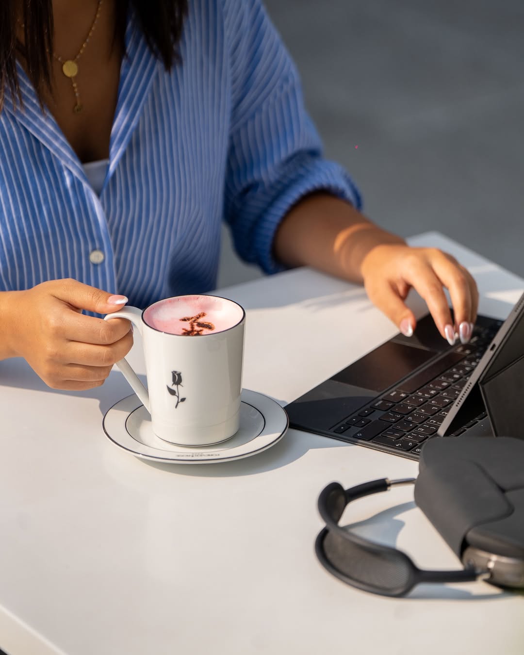 A person in a blue shirt works on a laptop next to a pink latte in a forever rose cafe mug.