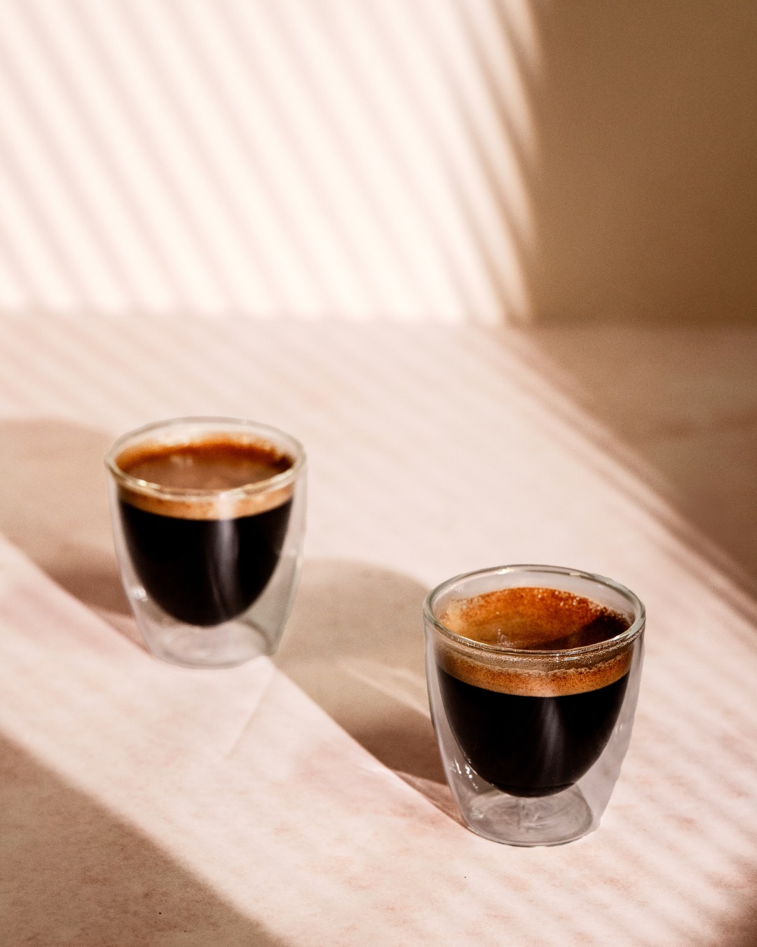 Two small glass cups of espresso on a light surface with soft, slatted window shadows.