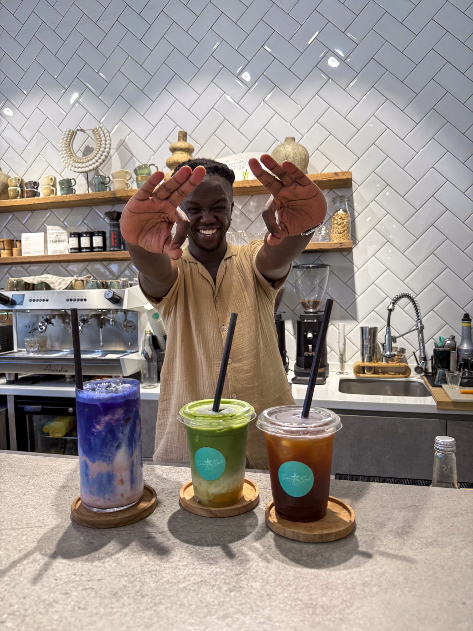 A smiling barista showing hand signs behind three colorful iced beverages on a cafe counter.