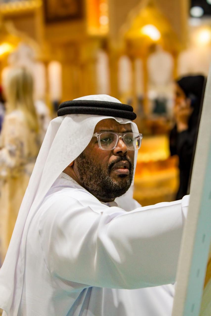 A man in traditional Emirati attire and glasses focused on a canvas in a brightly lit room.