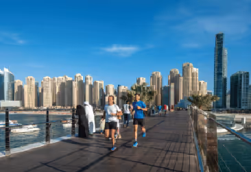 People jogging on a pedestrian bridge with the Dubai Marina skyline in the background.
