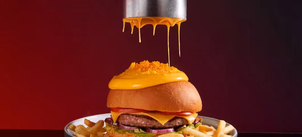 Molten cheese pouring onto a gourmet burger with fries on a dark red background.