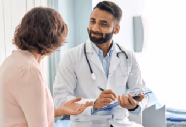 A male doctor in a white coat listens to a patient while holding a clipboard.