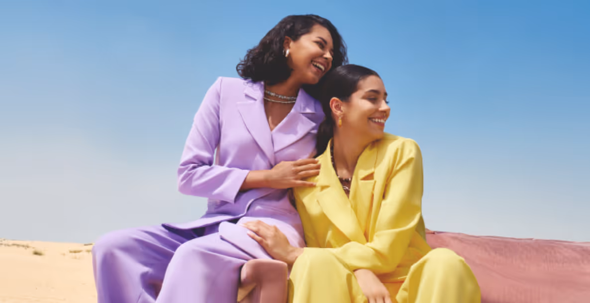 Two smiling women in vibrant purple and yellow suits sitting together in a desert setting.