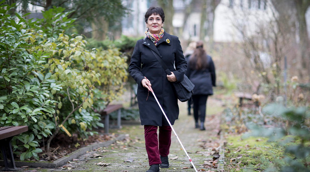 A blind woman walking through a park with a white cane to help her navigate.