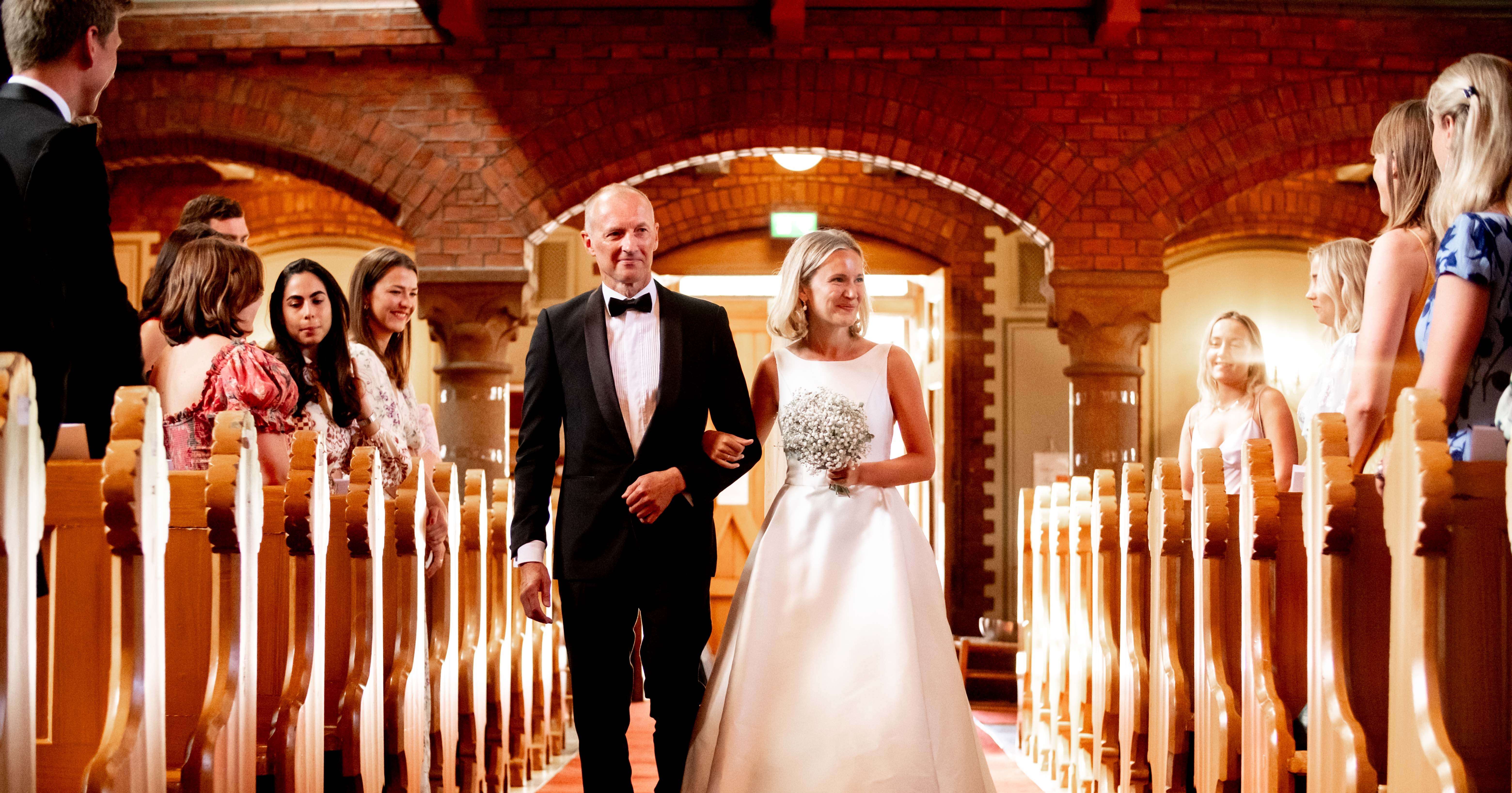 Bride in white gown holding flowers walking arm-in-arm down the church aisle with a man in a black tuxedo, surrounded by seated guests.