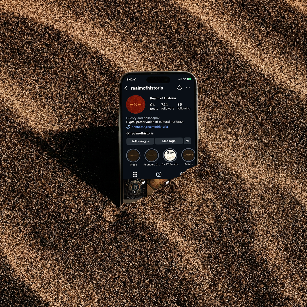 Smartphone partially buried in sand displaying Instagram profile 'realmofhistoria' focused on digital preservation of cultural heritage.