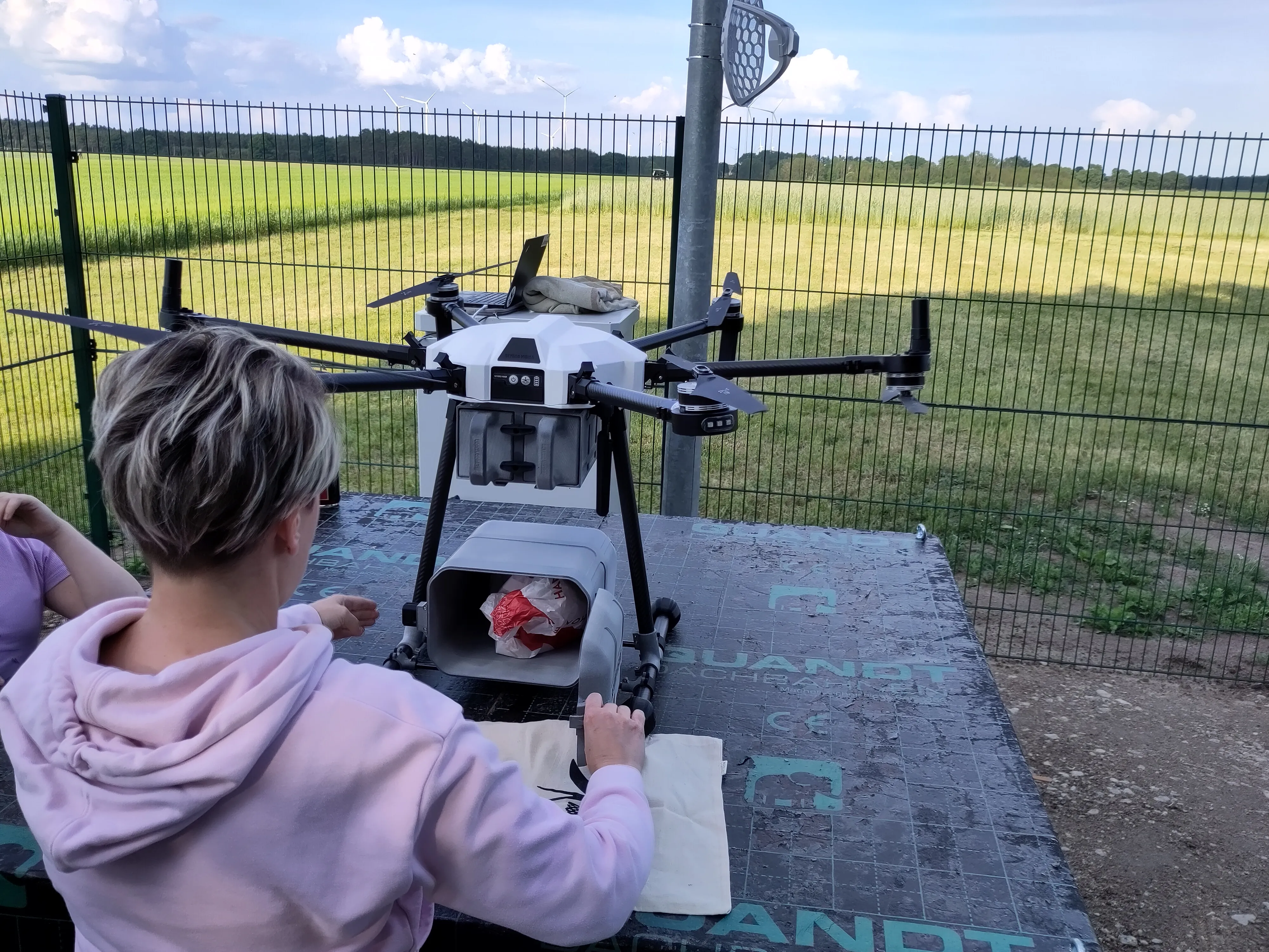 Frau in rosa Kapuzenpullover vor einer Marktschwalbe-Drohne mit offener Frachtbox auf einem Tisch im Freien.