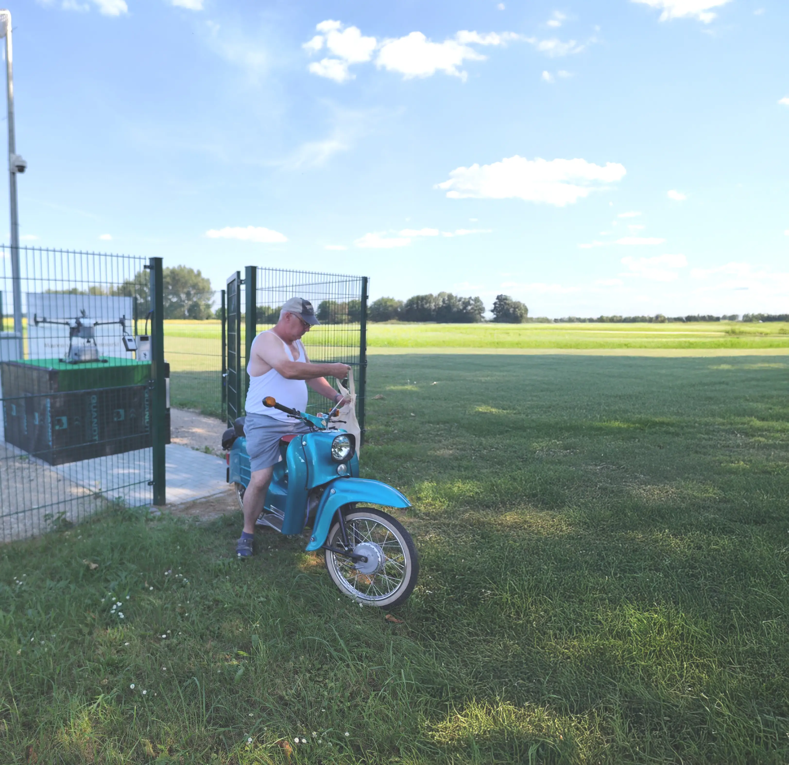 Mann in Shorts, Tanktop und Kappe steht neben einem blauen Simson-Moped auf grasbewachsenem Feld unter blauem Himmel, der gerade eine Lieferung von der Marktschwalbe abgeholt hat.