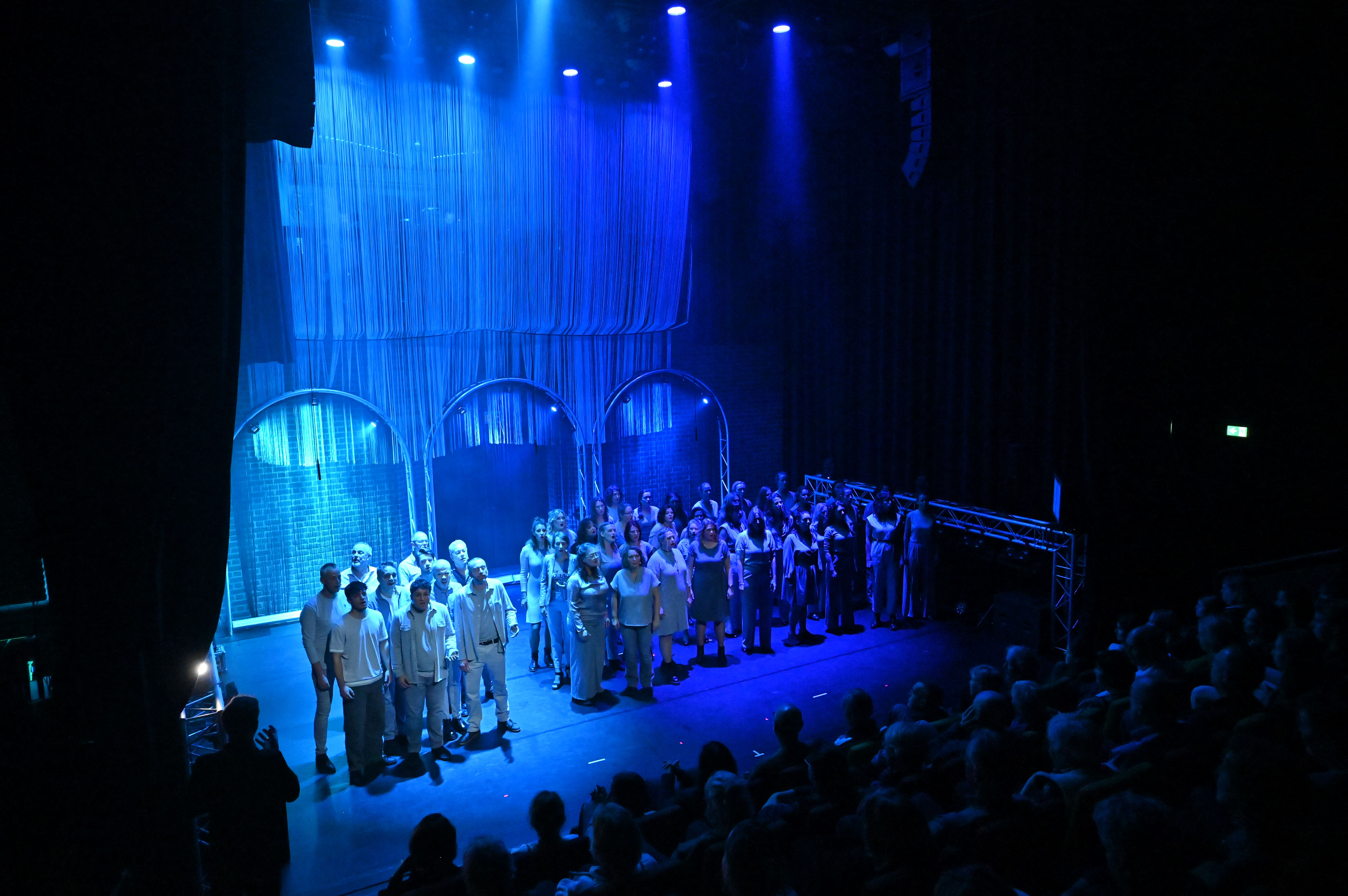 Volle zaal met een koor op een blauw uitgelicht podium