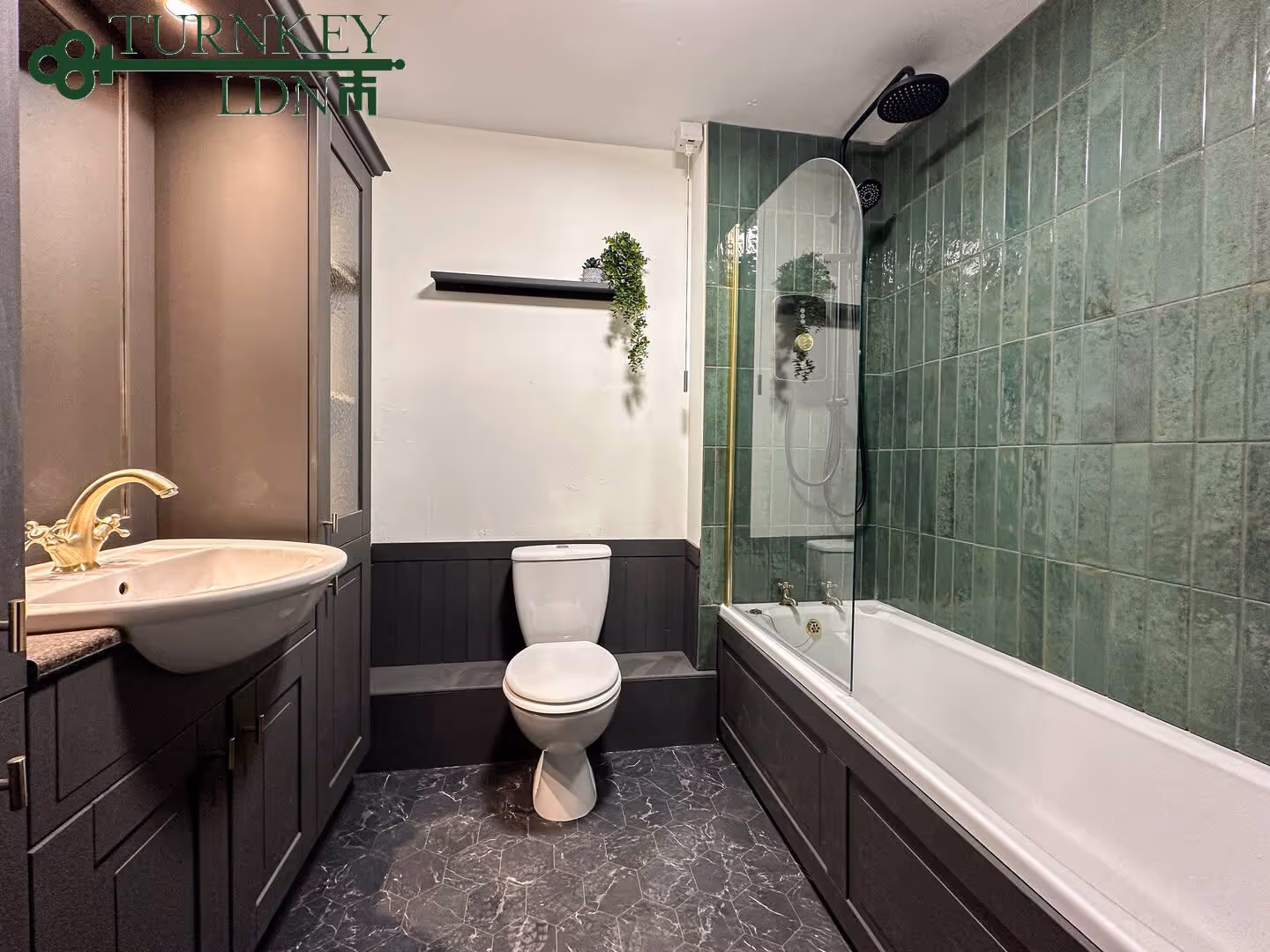 Modern bathroom with black cabinetry, white sink and toilet, green tiled wall around bathtub with black shower fixtures, and a small plant on a black wall shelf.