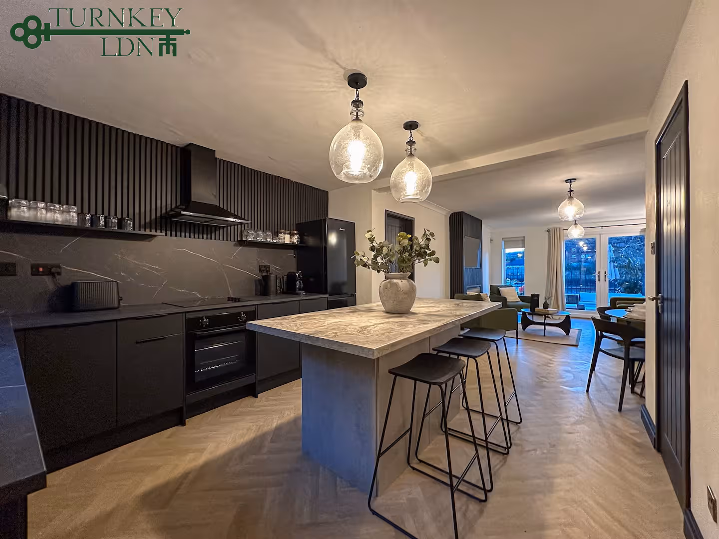 Modern kitchen with black cabinets, marble island with bar stools, pendant lights, and open living area with large windows.