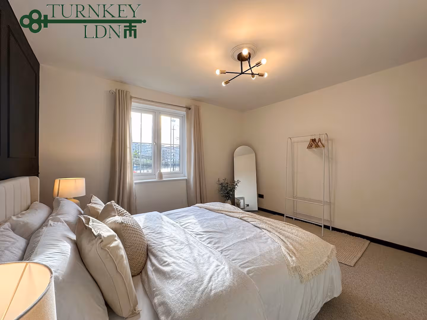 Cozy bedroom with a large bed, multiple pillows, a beige blanket, a standing mirror, and a clothes rack with hangers.