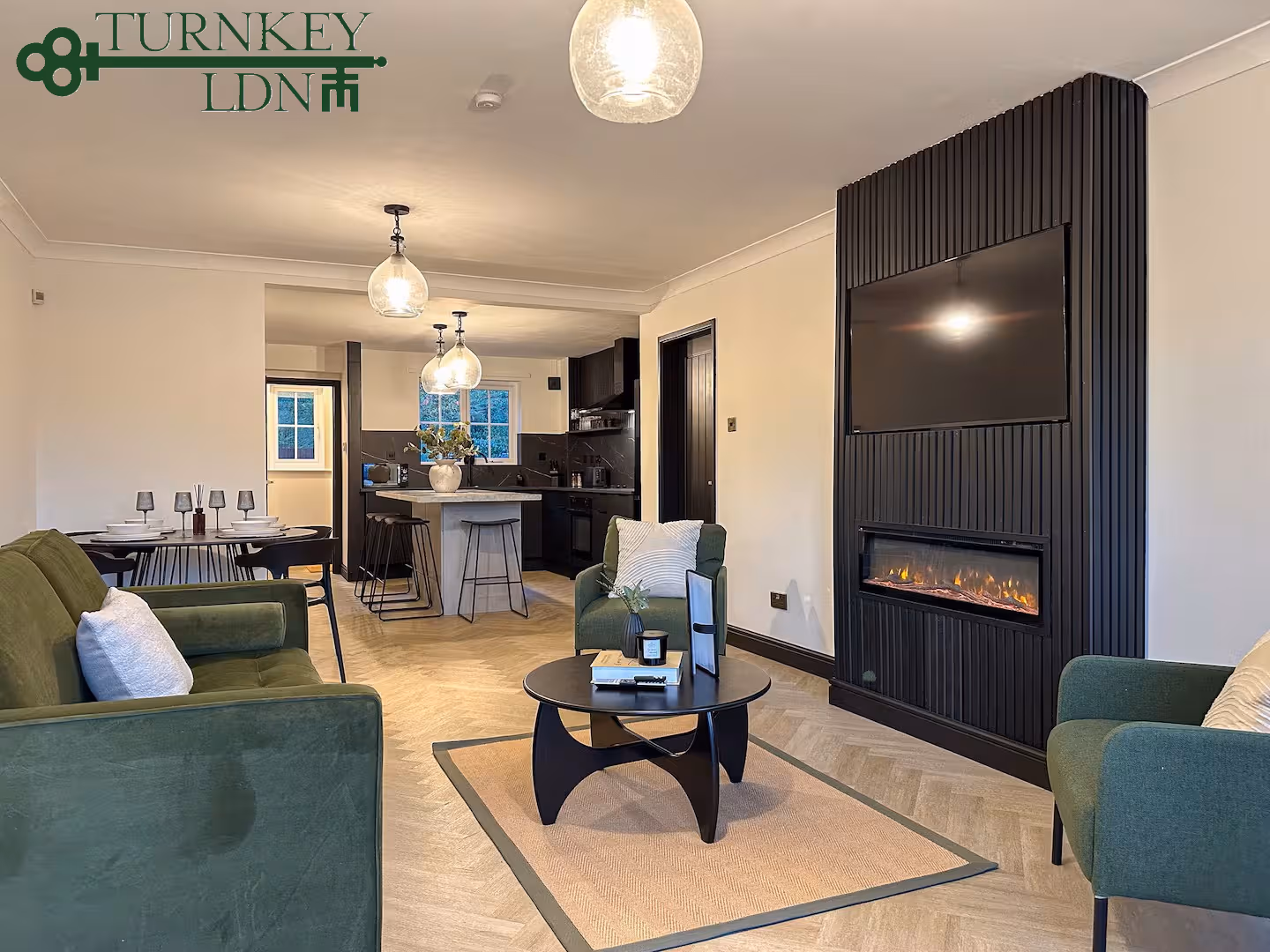 Modern living room with green velvet sofas and armchairs, black coffee table on beige rug, wall-mounted TV above a fireplace, and an open kitchen in the background with pendant lights.