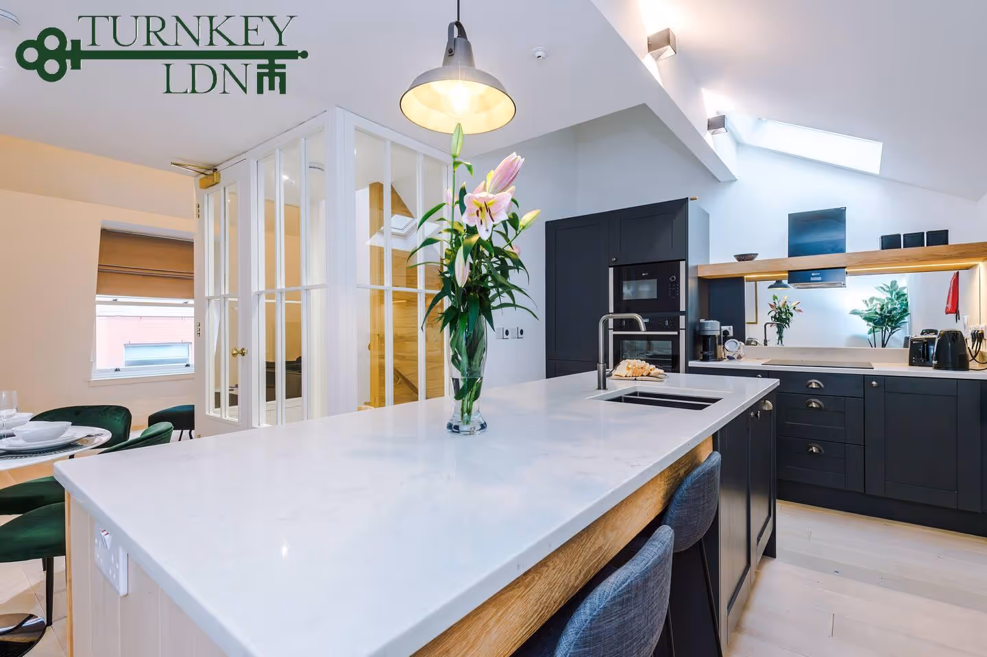 Modern kitchen with a large white marble island, dark cabinetry, stainless steel appliances, and a vase of pink lilies under a hanging lamp.