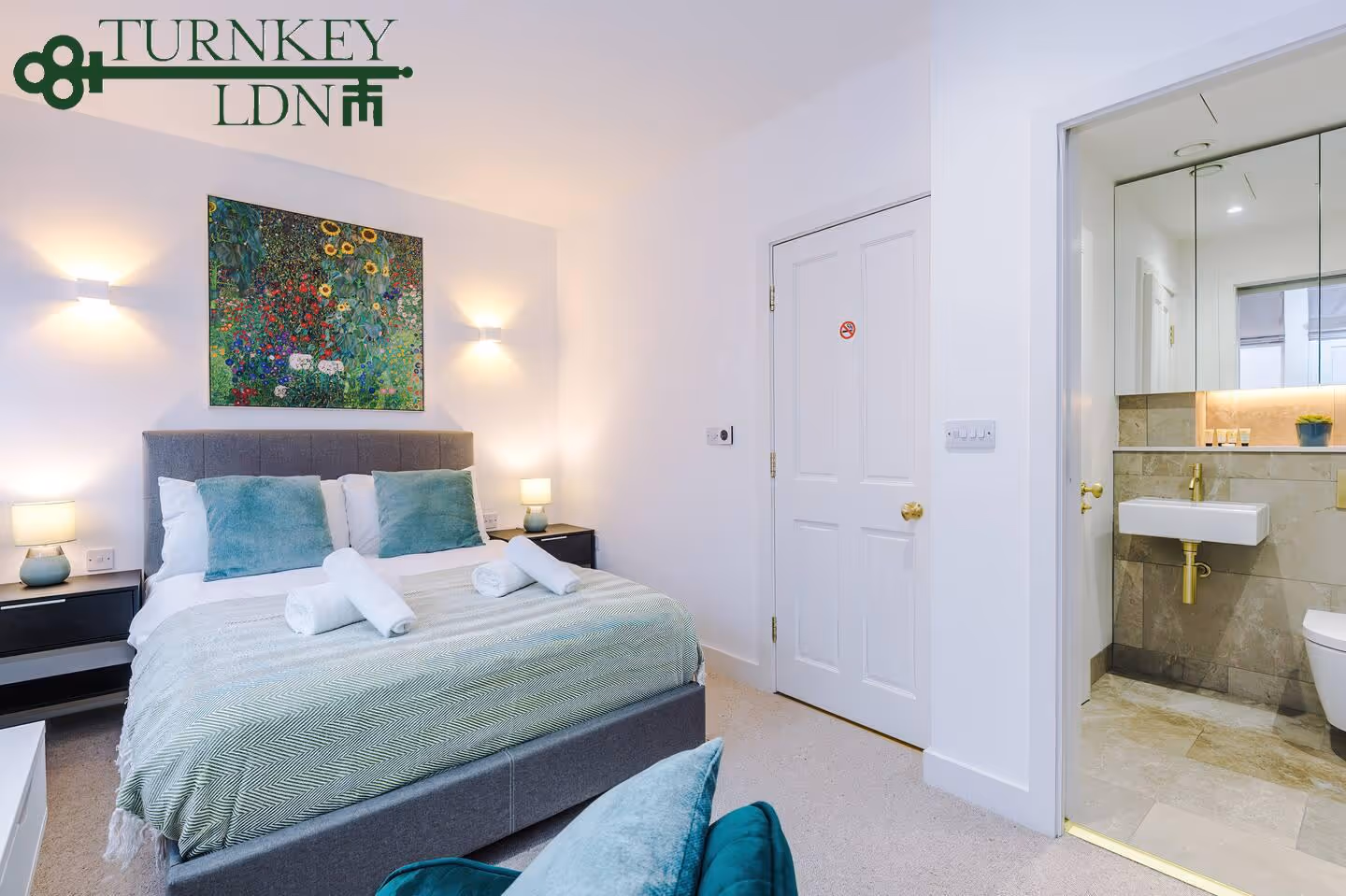 Modern bedroom with a gray upholstered bed, teal pillows, rolled white towels, floral painting above, and a view into a bathroom with a white sink and gold fixtures.