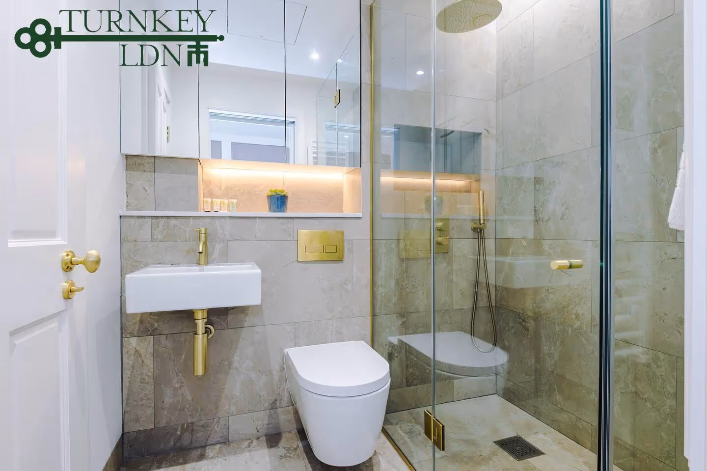 Modern bathroom with beige marble tiles, white sink and toilet, gold fixtures, and a glass-enclosed shower.