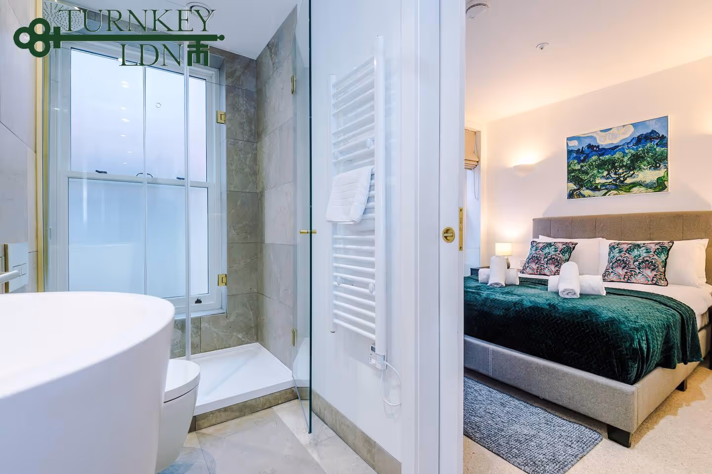 Modern bathroom with glass shower and white towel rack open to bedroom featuring bed with green bedspread and decorative pillows.