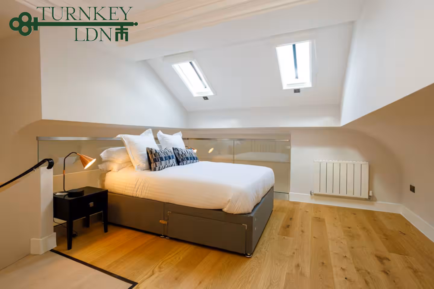 Modern attic bedroom with a double bed, white bedding, two skylights, wooden floor, and a small black bedside table with a lamp.