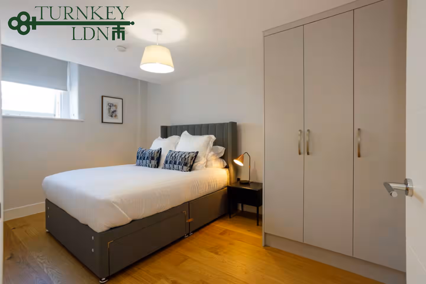 Minimalist bedroom with a made bed featuring white linens, gray upholstered headboard, black nightstand with a lamp, and a three-door wardrobe on hardwood floor.