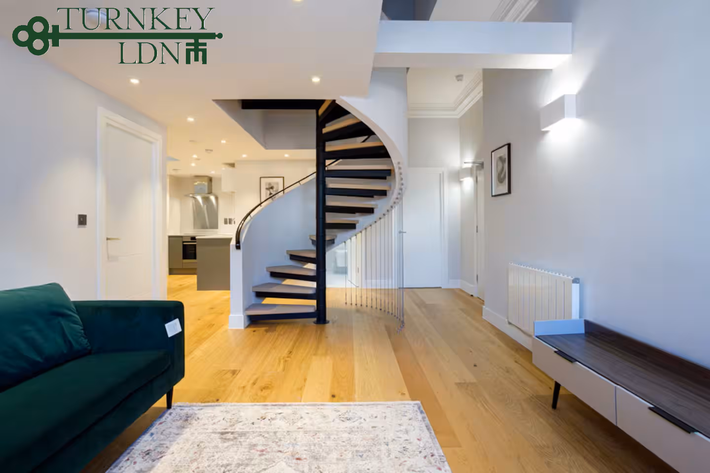 Modern living area with wooden flooring, a green sofa, a light patterned rug, and a black spiral staircase leading upstairs.