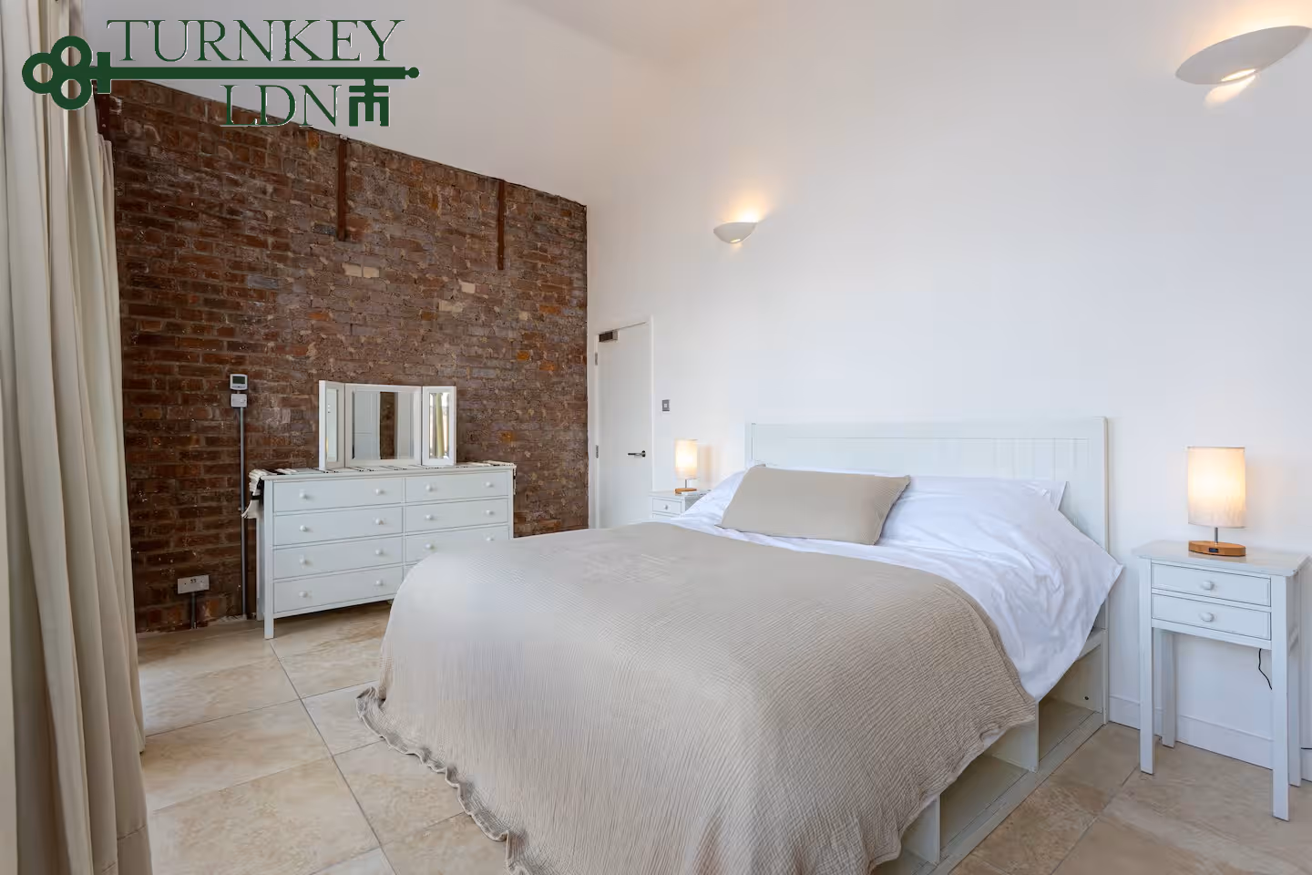 Minimalist bedroom with a large bed covered in beige linens, white bedside tables with lamps, exposed brick wall, and white dresser with mirror.