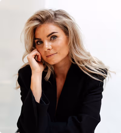 Vrouw met lang blond haar in een zwarte jas, leunend met haar hoofd op haar hand tegen een witte achtergrond.