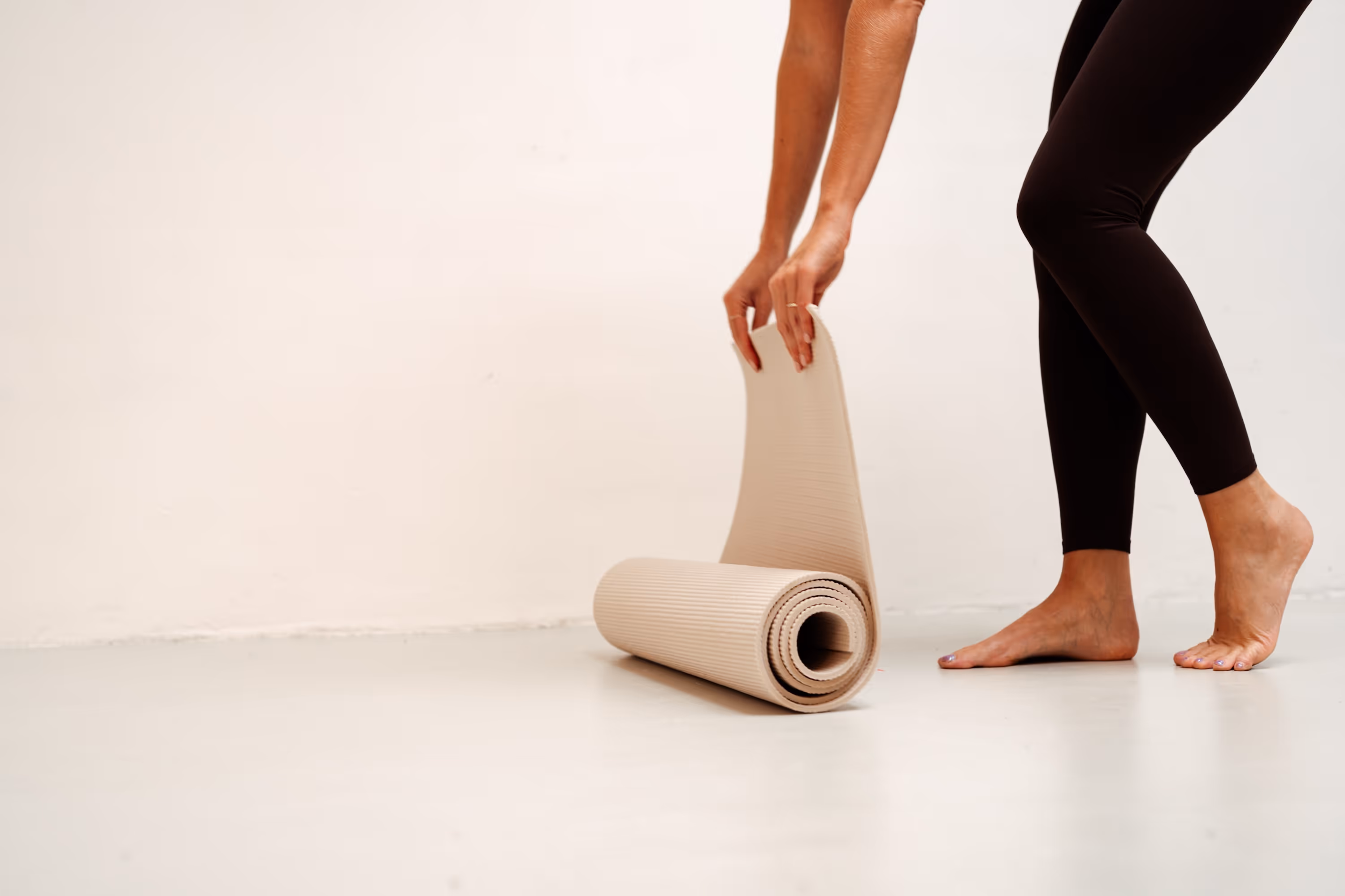 Persoon met zwarte legging begint een beige yogamat uit te rollen op een lichte vloer.