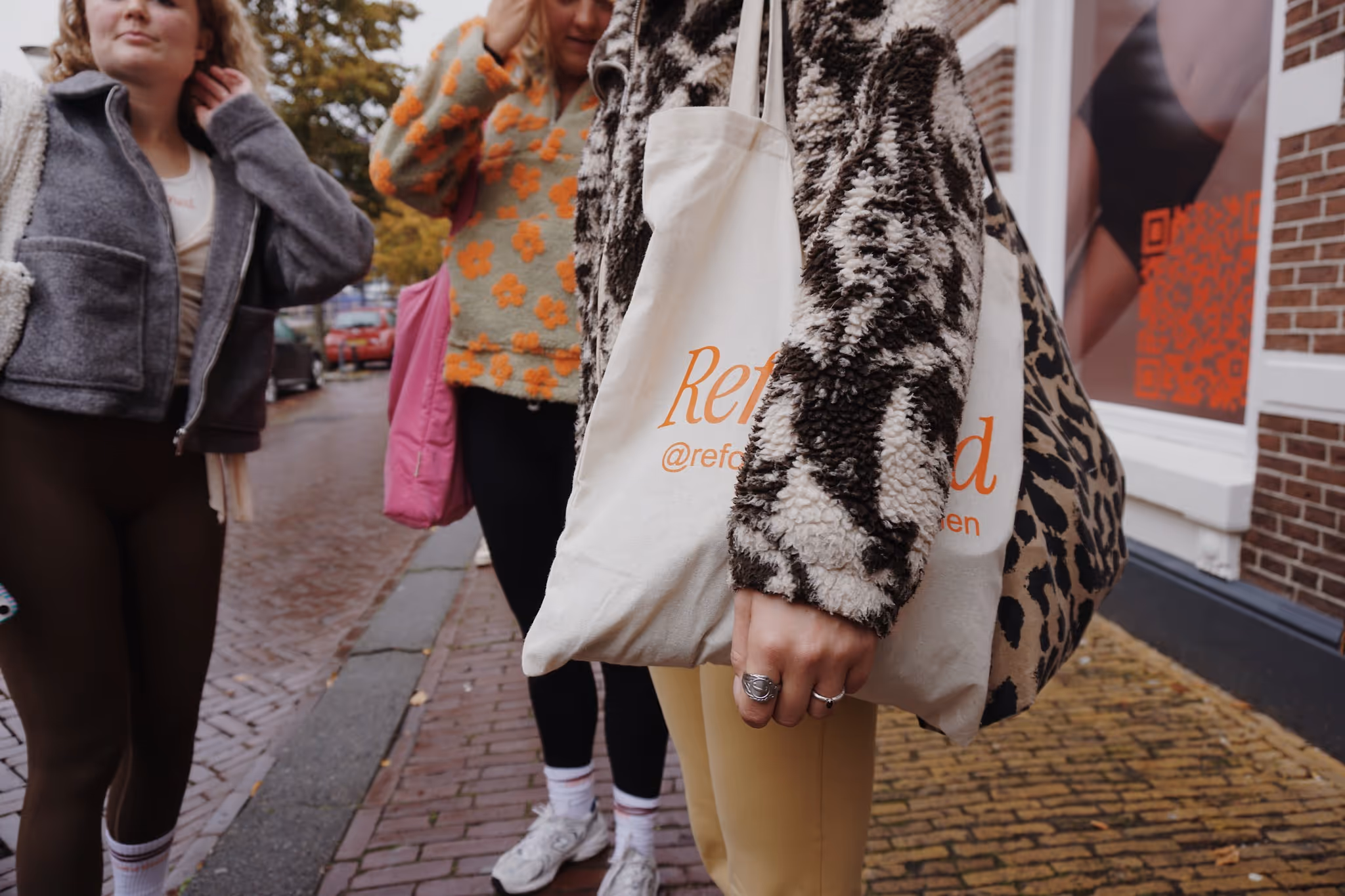 Drie mensen wandelen buiten op een straat met natte stenen, één draagt een witte tas met oranje tekst en een warm patroonjack.