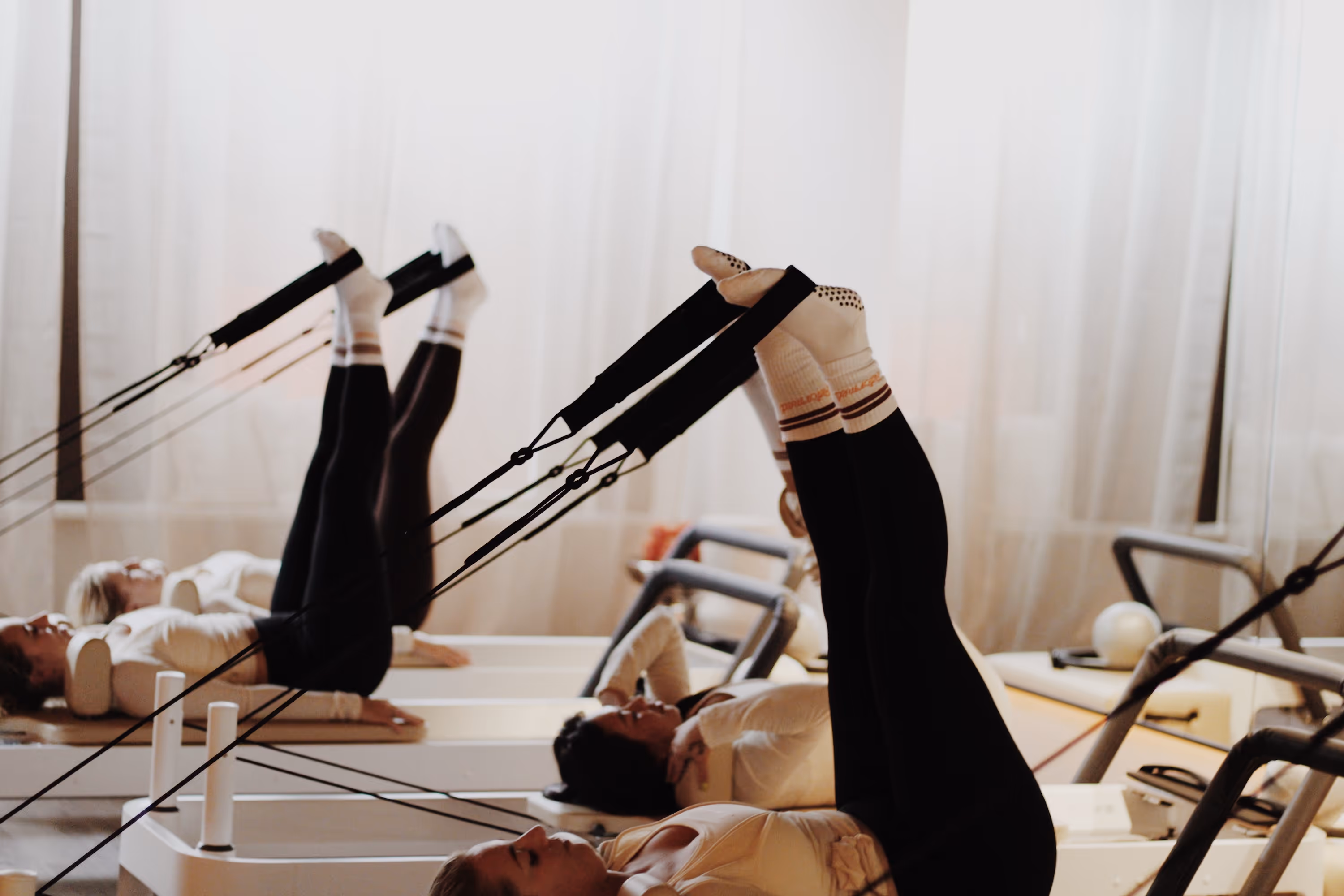 Groep mensen doet reformed pilates met benen omhoog en banden aan voeten in een studio.